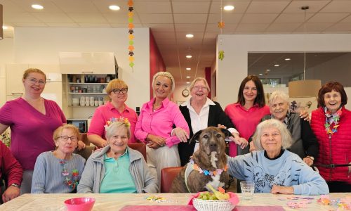 Gruppenfoto mit Pflege-Team, Seniorinnen und Senioren sowie Therapiehund im hellen Aufenthaltsraum.